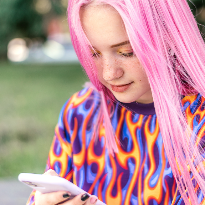 Junges Mädchen mit pinken Haaren und orange-blauem Pullover schaut auf ein Smartphone.