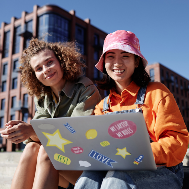 Zwei Mädchen sitzen auf Stufen vor einem modernen Bürogebäude. Auf dem Schoß des rechten Mädchens ist ein aufgeklappter Laptop mit Stickern.