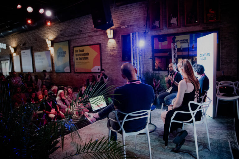 Das Bild zeigt eine Paneldiskussion im German Haus während der SXSW. Vier Personen sitzen auf einer Bühne vor einem Publikum. Die Atmosphäre ist lebhaft, mit bunten Lichtern und dekorativen Pflanzen.