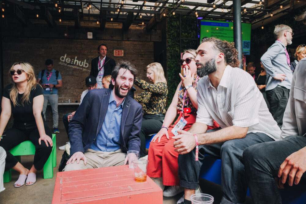 Eine Gruppe von Menschen sitzt in einer entspannten Atmosphäre zusammen. Sie lachen und unterhalten sich. Im Hintergrund sind weitere Teilnehmer und ein Schild mit der Aufschrift 'de:hub' zu sehen.