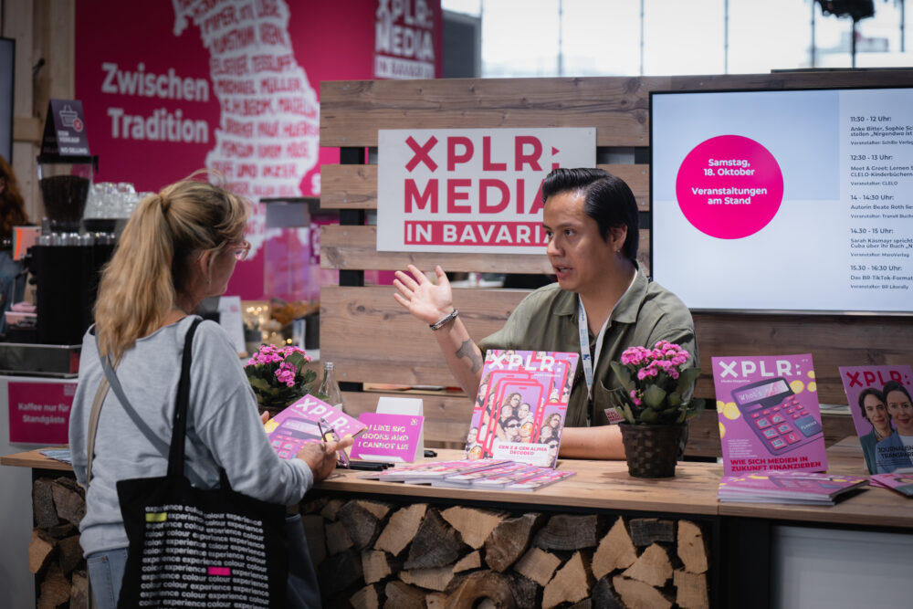 Ein Messestand von XPLR: MEDIA in Bavaria mit Holzdekor, Magazinen, Blumen und einer digitalen Anzeige. Eine Person informiert eine Besucherin über die ausgestellten Inhalte und Veranstaltungen.