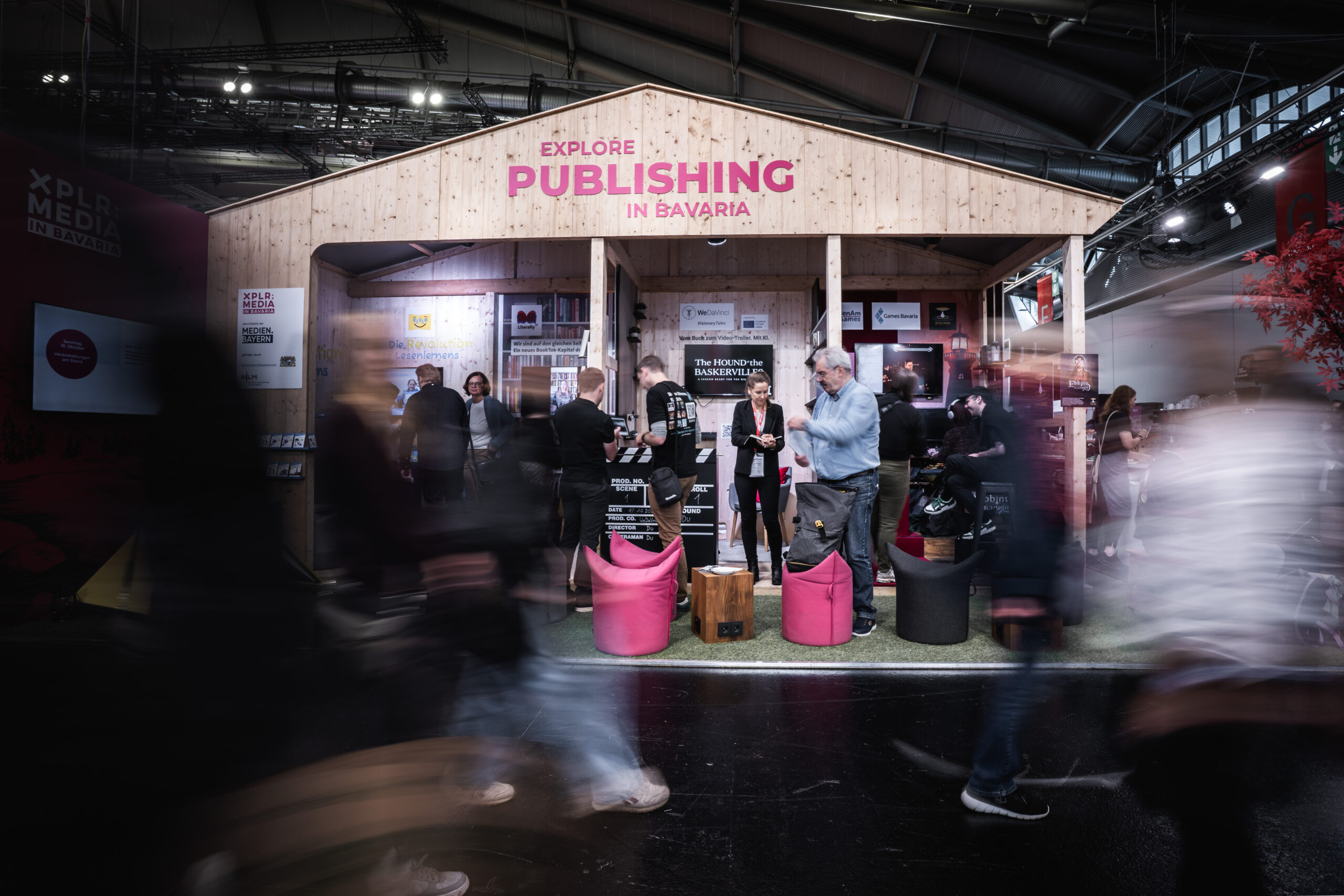 Ein Messestand aus Holz mit der Aufschrift 'Explore Publishing in Bavaria' präsentiert bayerische Verlage. Menschen stehen und unterhalten sich, während andere vorbeigehen. Im Hintergrund sind Bücher und Poster zu sehen.
