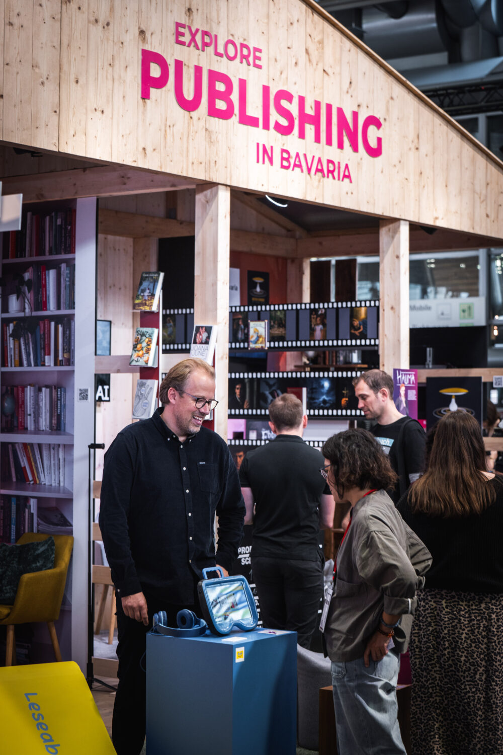 Ein Messestand mit Holzoptik präsentiert unter dem Motto 'Explore Publishing in Bavaria' Bücher und Projekte bayerischer Verlage. Menschen stehen im Gespräch, während Bücher und Medien ausgestellt sind. Im Vordergrund ein blauer Tisch mit Kopfhörern und einem Gerät.