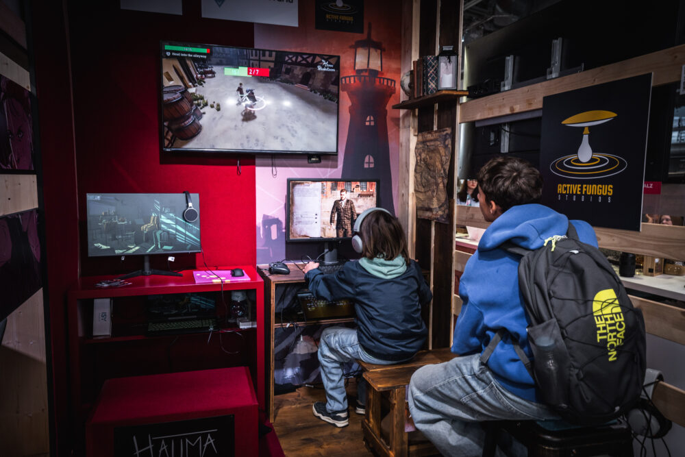 Zwei Personen sitzen an einem Stand mit Gaming-Setup. Auf Monitoren laufen Videospiele, darunter ein Actionspiel und ein Detektivspiel. Der Stand ist mit roten und holzfarbenen Elementen gestaltet, Poster und Logos wie 'Active Fungus Studios' sind sichtbar.