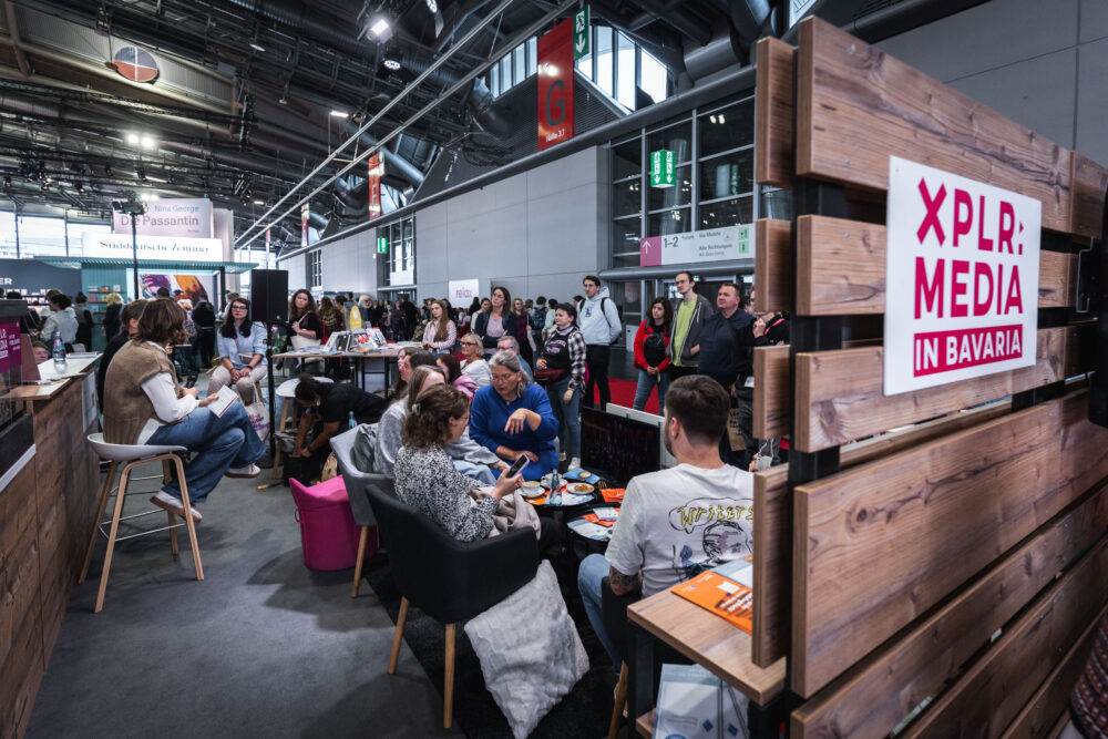 Ein Messestand von XPLR: MEDIA in Bavaria auf der Frankfurter Buchmesse. Menschen sitzen in einer gemütlichen Lounge, lesen, unterhalten sich und präsentieren Projekte. Im Hintergrund sind weitere Stände und Besucher der Messe zu sehen.