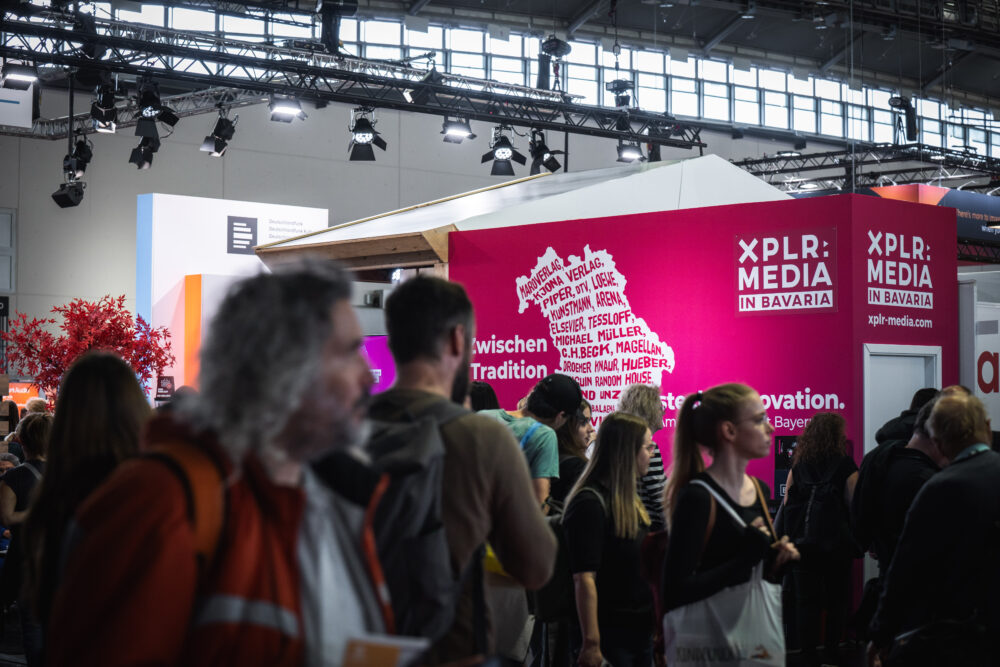 Ein Messestand mit pinkem Hintergrund und der Aufschrift 'XPLR: MEDIA in Bavaria'. Menschen stehen davor, während bayerische Verlage und Projekte präsentiert werden. Im Hintergrund sind Lichter und ein roter Baum zu sehen.