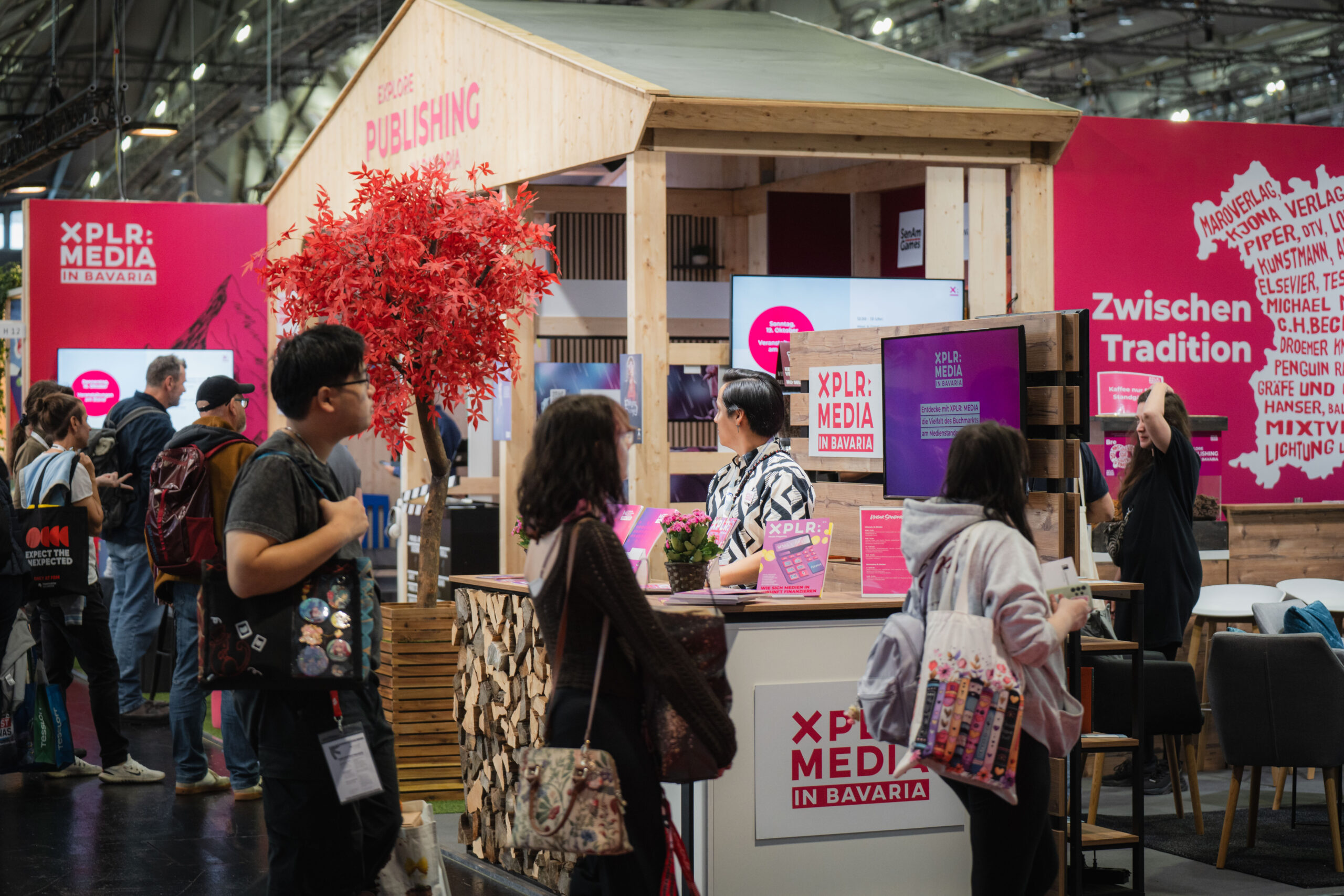 Ein Messestand von XPLR: MEDIA in Bavaria mit Holzdesign, roten Akzenten und einem roten Baum. Menschen informieren sich über Projekte, während Broschüren und Bildschirme präsentiert werden. Im Hintergrund sind weitere Stände und leuchtende Schilder zu sehen.